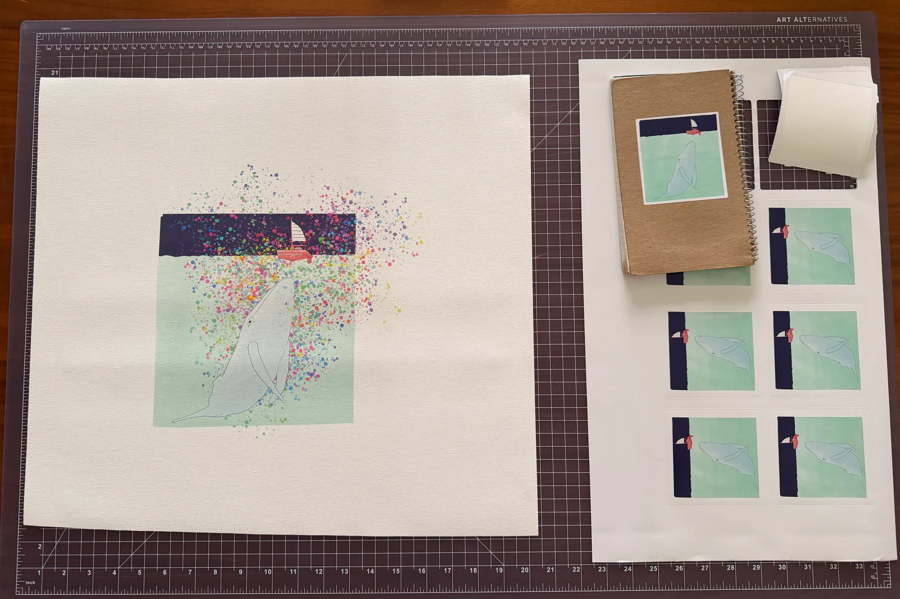 The printed whale artwork with rainbow brainbows on canvas, next to sheets of kiss-cut stickers and a kraft notebook, laid out on a cutting mat.