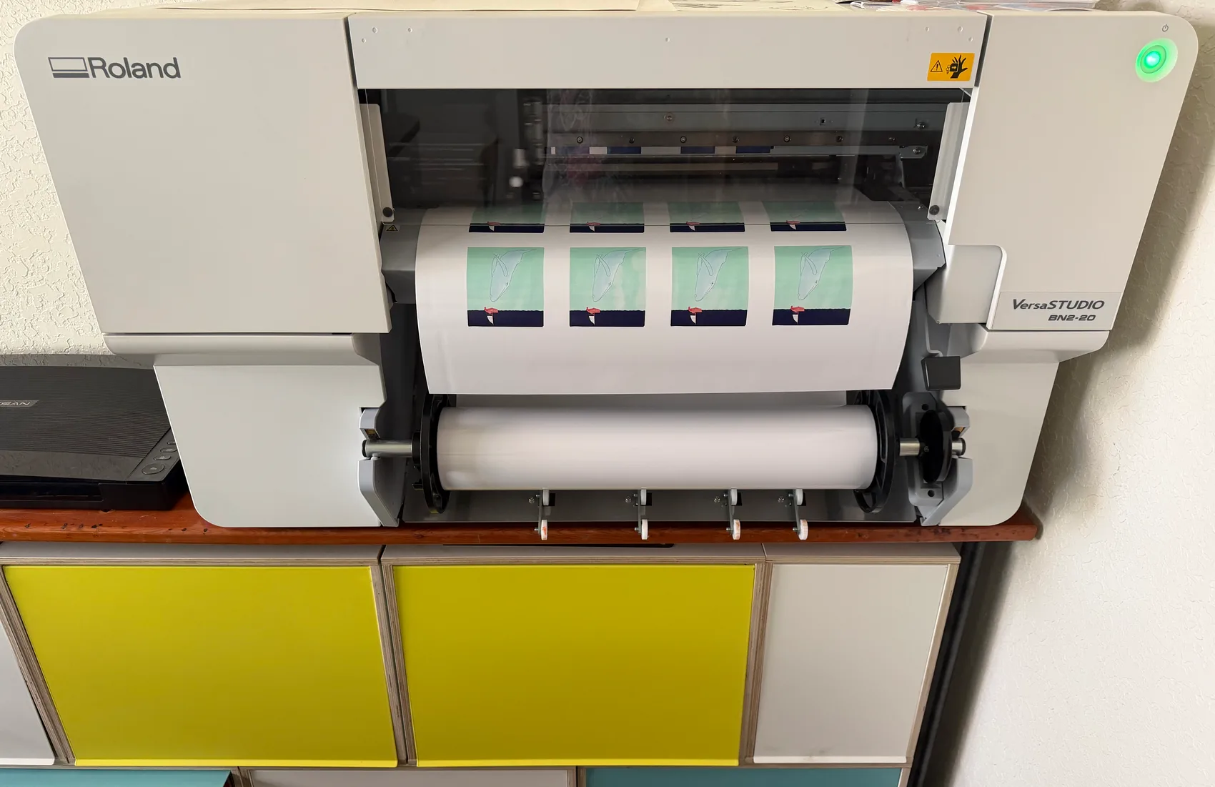 A Roland VersaSTUDIO print-and-cut machine running a roll of the printed whale stickers, colorful shelving below.